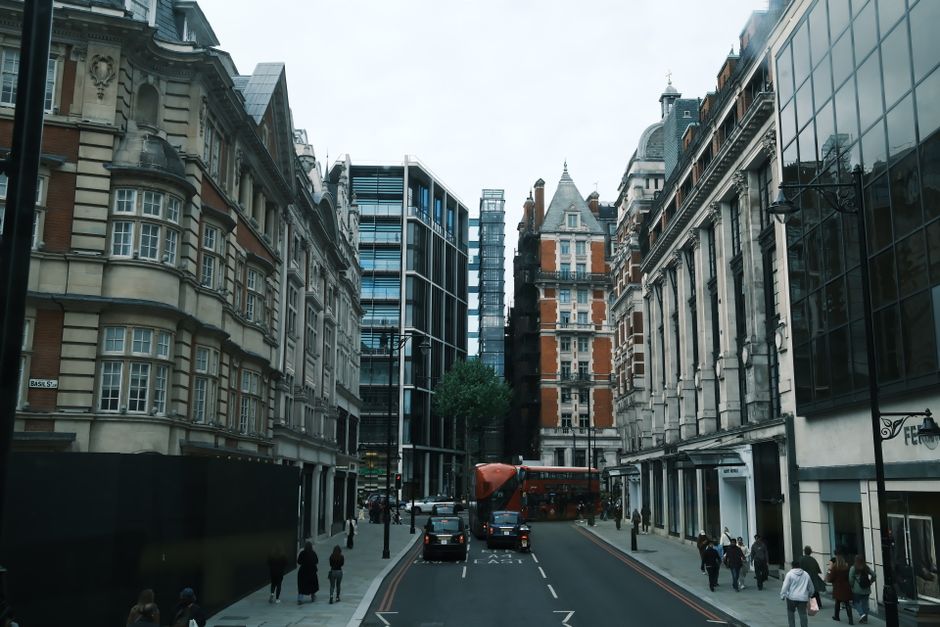 A large green box trailer is parked on a city street in front of modern high-rise buildings with glass facades and older stone structures. The trailer is attached to a vehicle out of view and is posit
