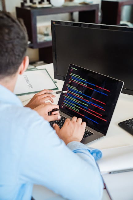 A person wearing a light blue shirt is seated at a desk, working on a laptop that displays lines of colorful computer code. The workspace includes a black monitor, a white document titled 'PERMISSION,